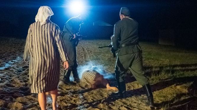 Aktorzy wcielają się tym razem w role więźniów i gestapowcow. Jeden z więźniów upadł, gestapowcy grożą mu bronią. Za nimi idzie kolejna więźniarka, w bialej chuście i pasiaku, ma gołe nogi.