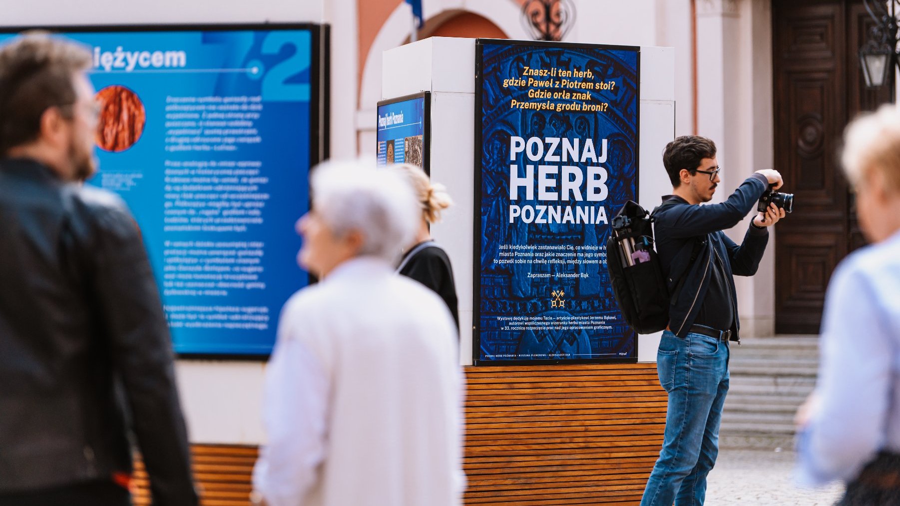 Ludzie oglądają plansze wystawy plenerowej, ktoś rozmawia, ktoś fotografuje,