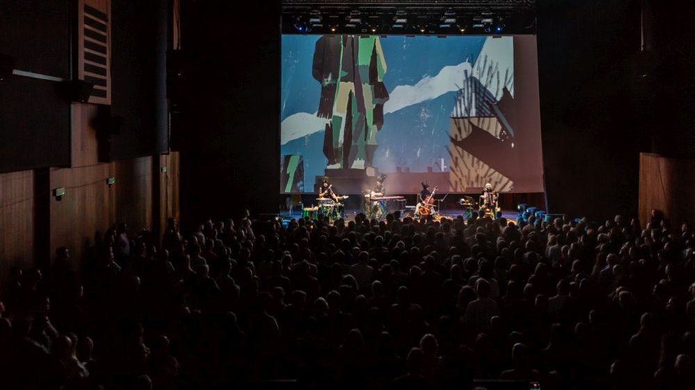 Tłum pod sceną. Na scenie trwa koncert zespołu z wizualizacjami w tle.