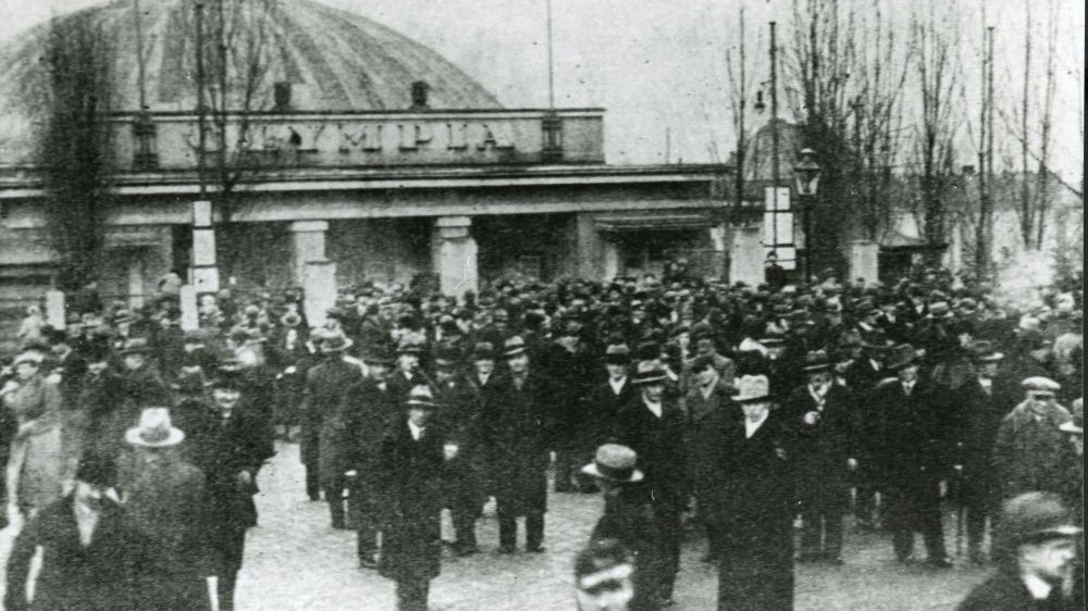 Tłum elegancko ubranych mężczyzn w płaszczach i kapeluszach na tle okrągłej budowli. Fotografia jest bardzo stara, niewyraźna.