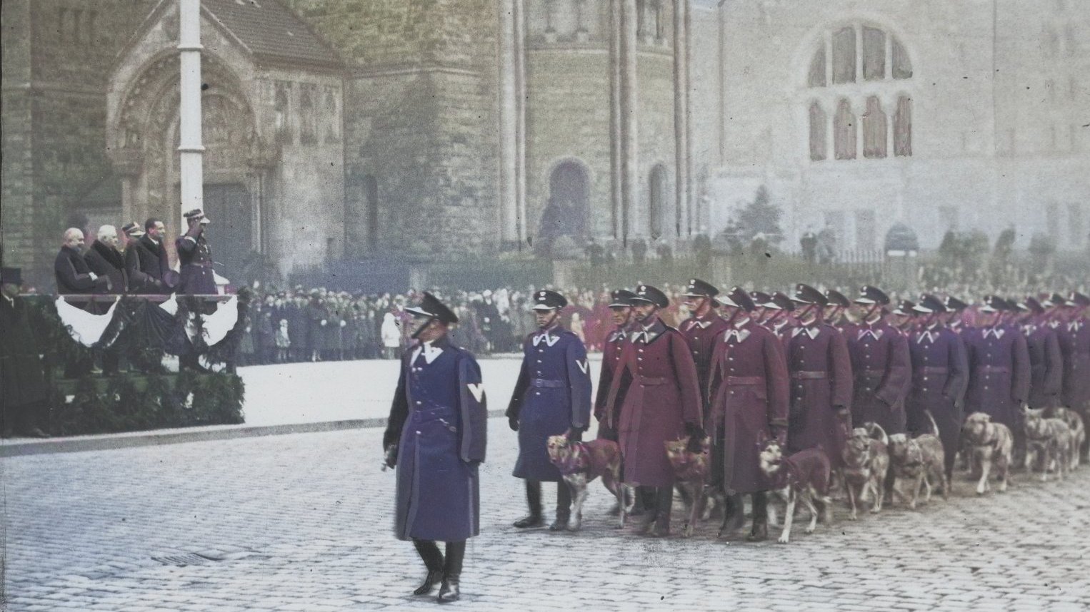 Umundurowani policjanci maszerują środkiem ybrukowanej ulicy z psami. Przyglądają im się ważne osobistości. W tle zabytkowe budynki. Czuć powagę.