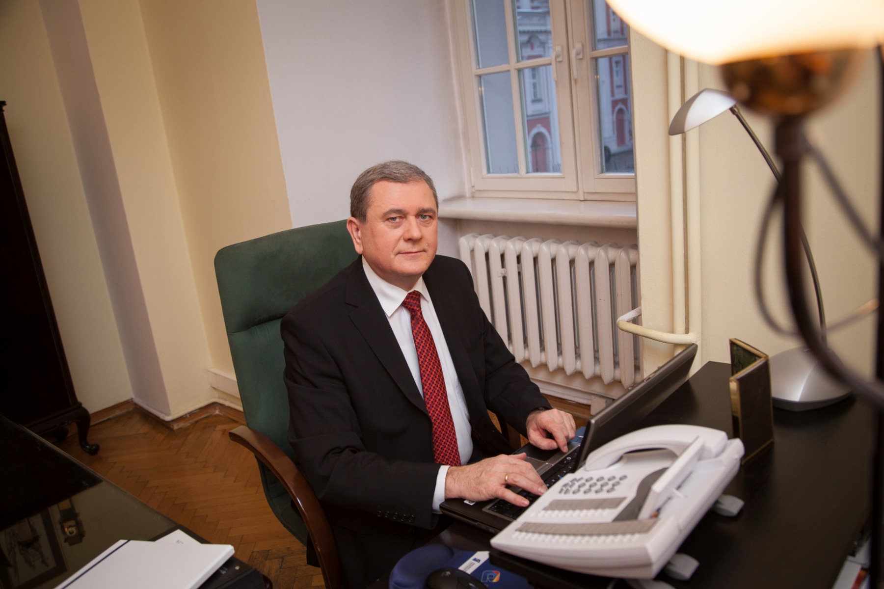 Picture of City Secretary. He is sitting at the desk with laptop. Men wearing a black suit and red tie.