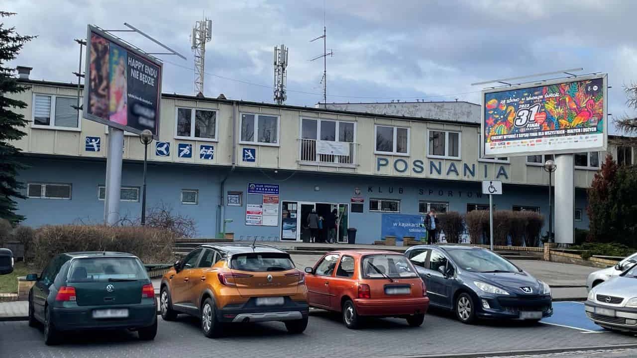 Front budynku Basenu Posnania. Budynek jest podłuższy i składa się z dwóch pięter. Pierwsze piętro jest niebieskie, druie żółte. Na piętrze napis "Posnania" i ikonografiki. Przed budynkiem stoją dwa bilboardy. Na parkinku przed budynkiem stoją zaparkowane samochody.