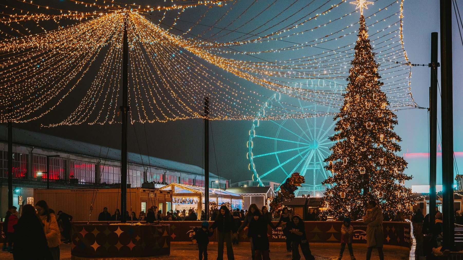 Widok na jarmark na targach. Na pierwszym planie widać grupkę osób na lodowisku. Po prawej duża choinka, w tle widać młyńskie koło. Po lewej u góry świąteczne iluminacje.