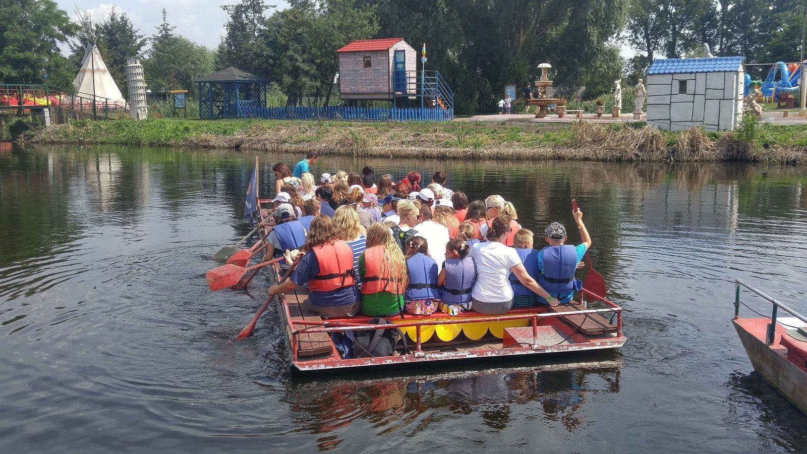 Uczestnicy wycieczki na tratwie. Na brzegu tipi, domki dla dzieci i plac zabaw.