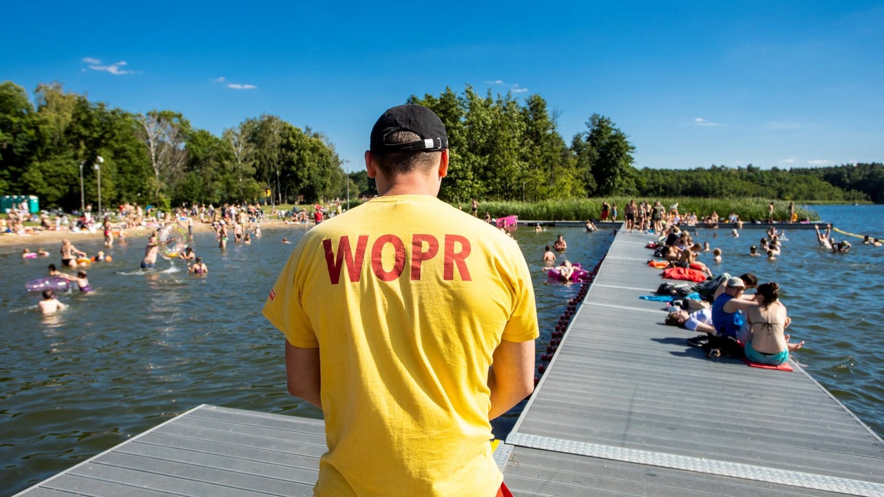 Zdjęcie przedstawia ratownika stojącego tyłem nad jeziorem. W wodzie, na pomoście i na plaży widać tłum ludzi.