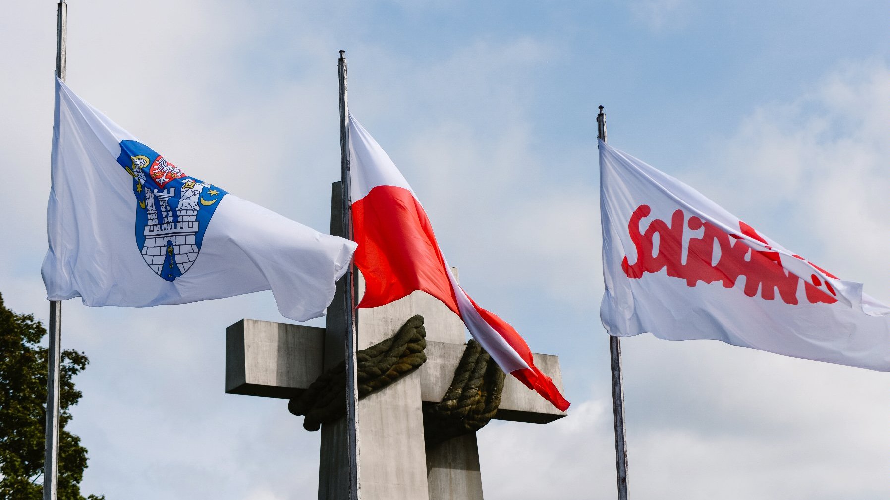 Zdjęcie przedstawia flagi Polski, z herbem Poznania i znakiem Solidarności. W tle znajduje się pomnik Dwóch Krzyży.