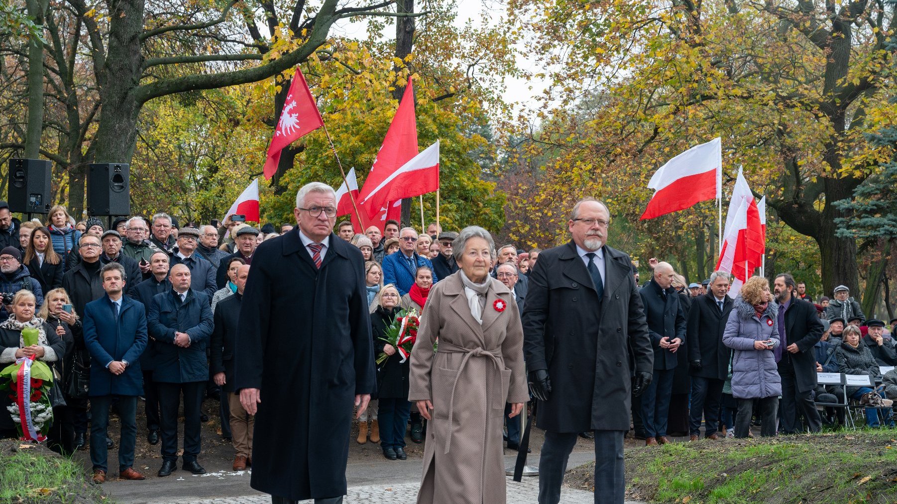 Na zdjeciu dwóch mężczyzn i kobieta, za nimi ludzie z flagami Polski