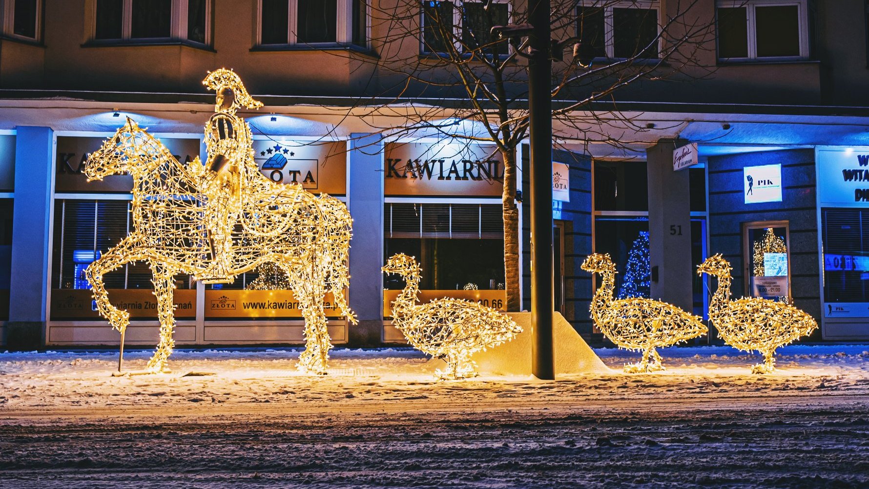 Zdjęcie przedstawia iluminację w kształcie Świętego Marcina na koniu oraz trzech idących za nim gęsi. Iluminacja znajduje się na ulicy. W tle widać witryny sklepów.