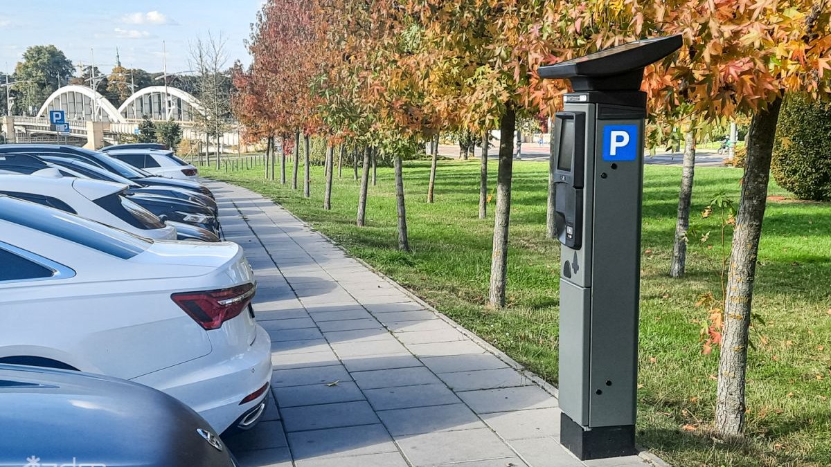 Zdjęcie parkomatu stojącego na chodniku pośród drzew z kolorowymi liśćmi. Po drugiej stronie chodnika rząd samochodów. W tle widac most św. Rocha