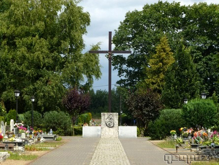 Na środku alejka. Po dwóch stronach groby. Na końcu alejki duży krzyż. Po dwóch stronach krzyża drzewa i krzewy.