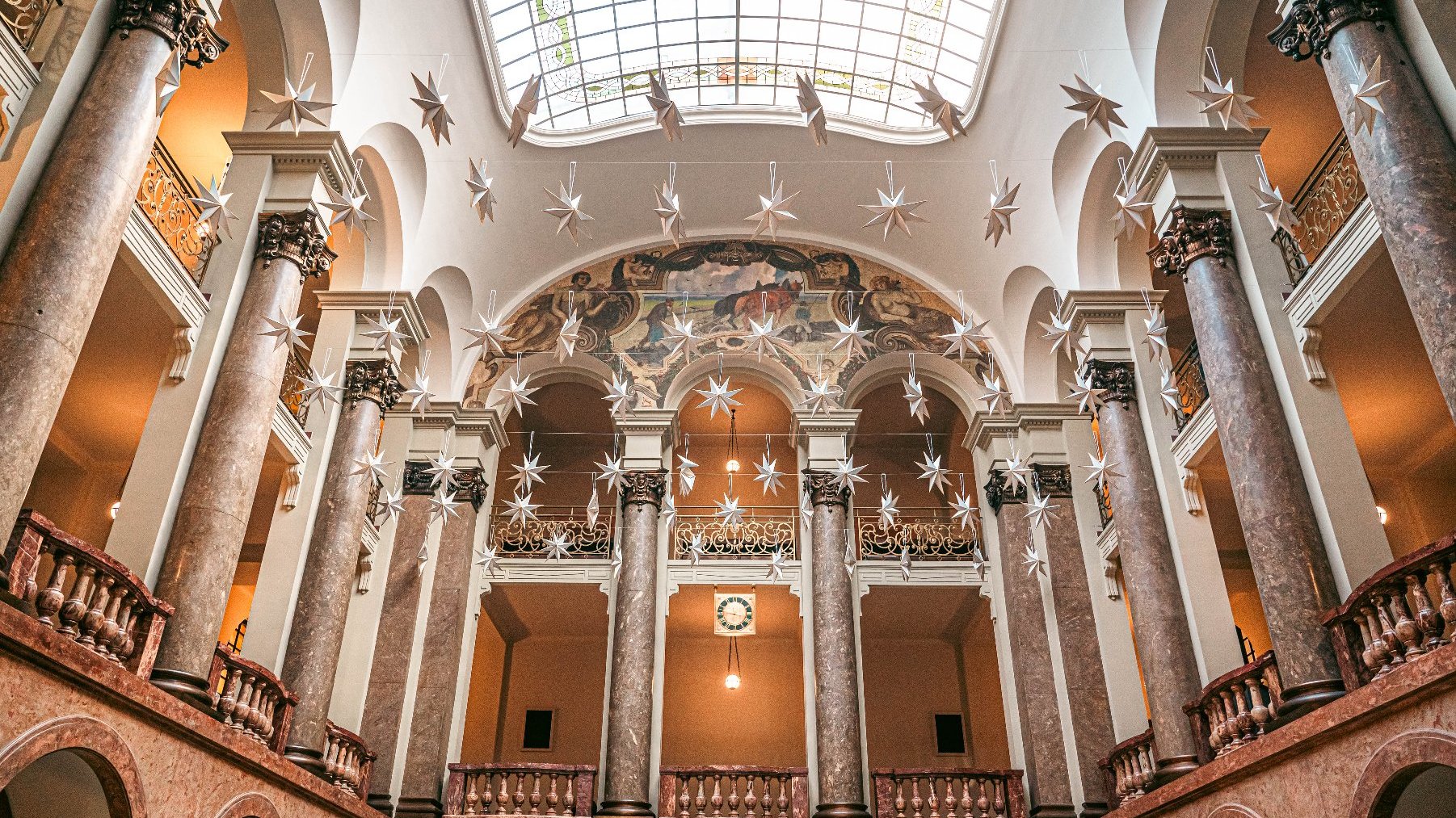 Wnętrze dużego, ozdobnego budynku z klasycznymi elementami architektonicznymi. Przestrzeń ma wysokie marmurowe kolumny z misternymi kapitelami, łukowe otwory i wysokie sklepienie z dużym świetlikiem. Sufit zdobią liczne gwiazdkowe dekoracje wiszące w dół. Na dalekiej ścianie, ponad łukami, widoczny jest mural, a w centrum obrazu znajduje się zegar. Budynek ma kilka poziomów z balkonami i balustradami.