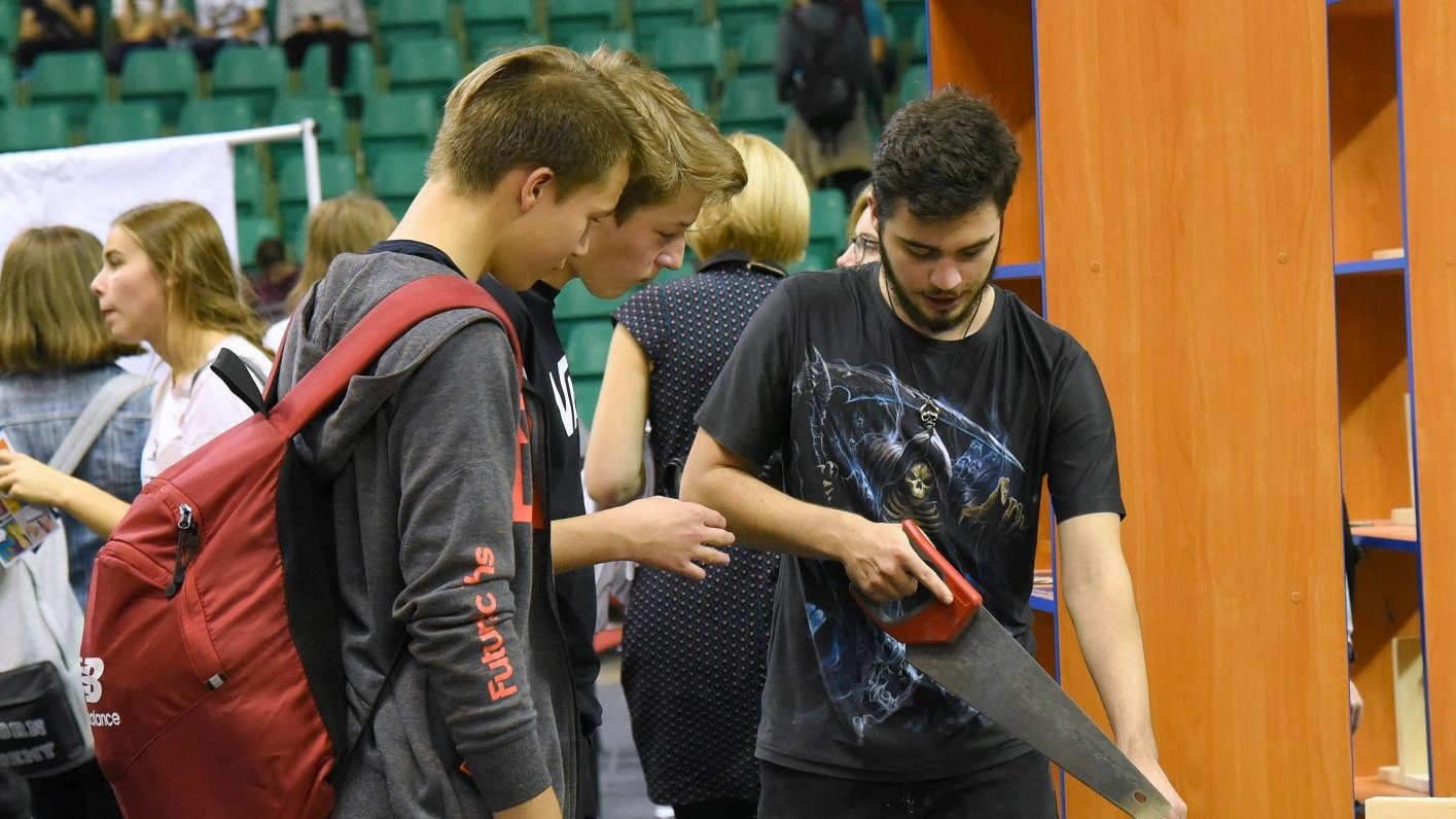 Galeria przedstawia zdjęcia z Areny Zawodów. Na pierwszym planie fotografii znajduje się trójka chłopców - jeden z nich prezentuje pozostałej dwójce piłę ręczną do cięcia drewna.