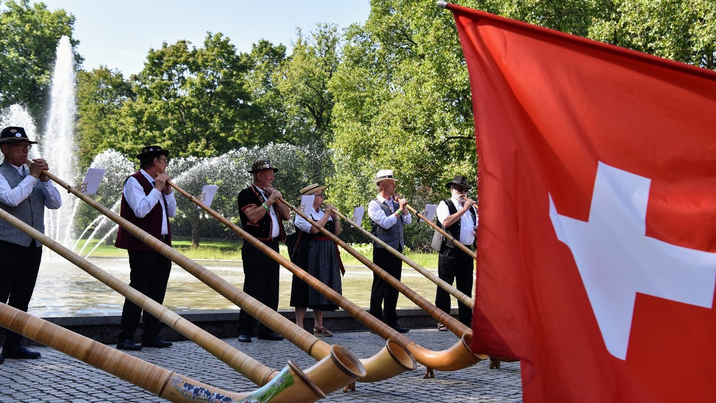 Na zdjęciu muzycy grający na rogach, w parku, przed nimi flaga Szwajcarii