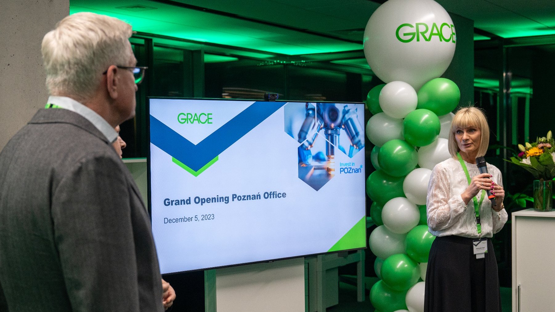 Zdjęcie przedstawia dwie osoby. Pomiędzy nimi widać telewizor, na którym widnieje grafika z napisem "Grace. Grand Opening Poznań Office".