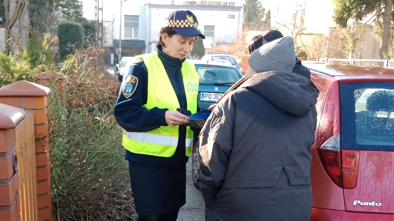 Zdjęcie przedstawia strażniczkę miejską na drodze, rozmawiającą z mieszkańcami.