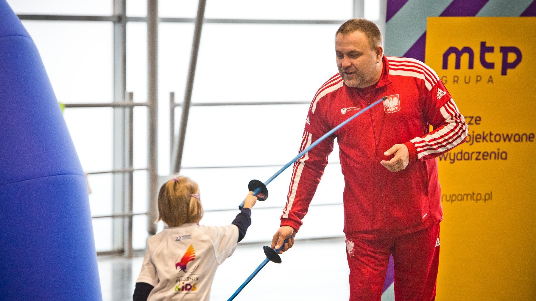 Zdjęcie przedstawia dziecko i dorosłego mężczyznę podczas szermierczego pojedynku.