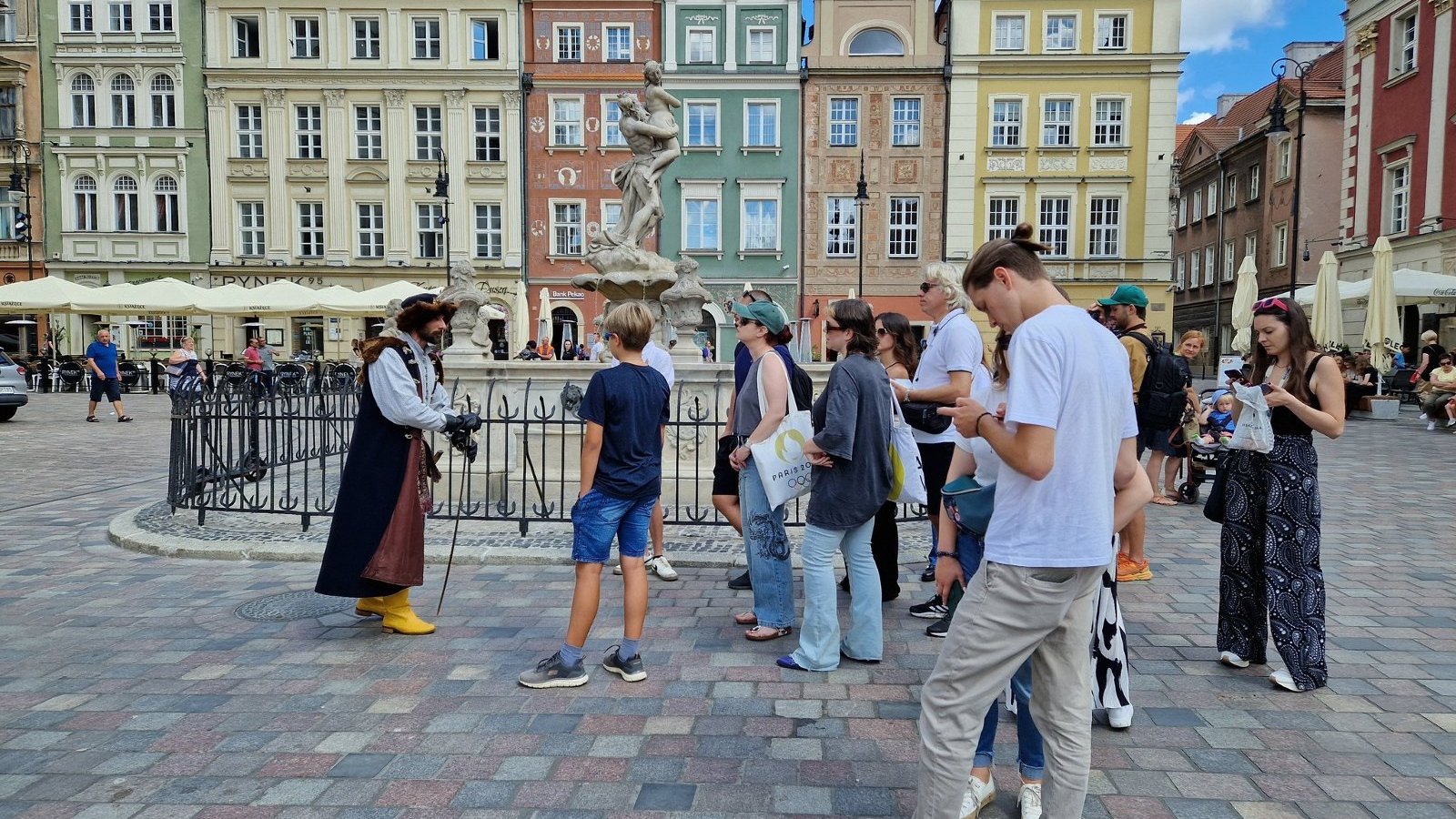 Na zdjęciu grupa ludzi na Starym Rynku, oprowadzana przez przewodnika w historycznym stroju