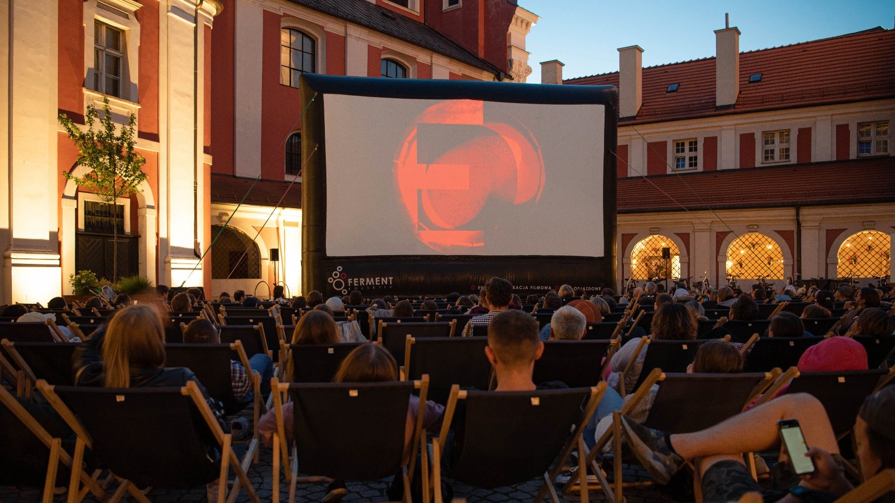 Zdjęcie przedstawia ludzi na leżakach oglądających film na telebimie na dziedzińcu urzędu miasta. - grafika artykułu