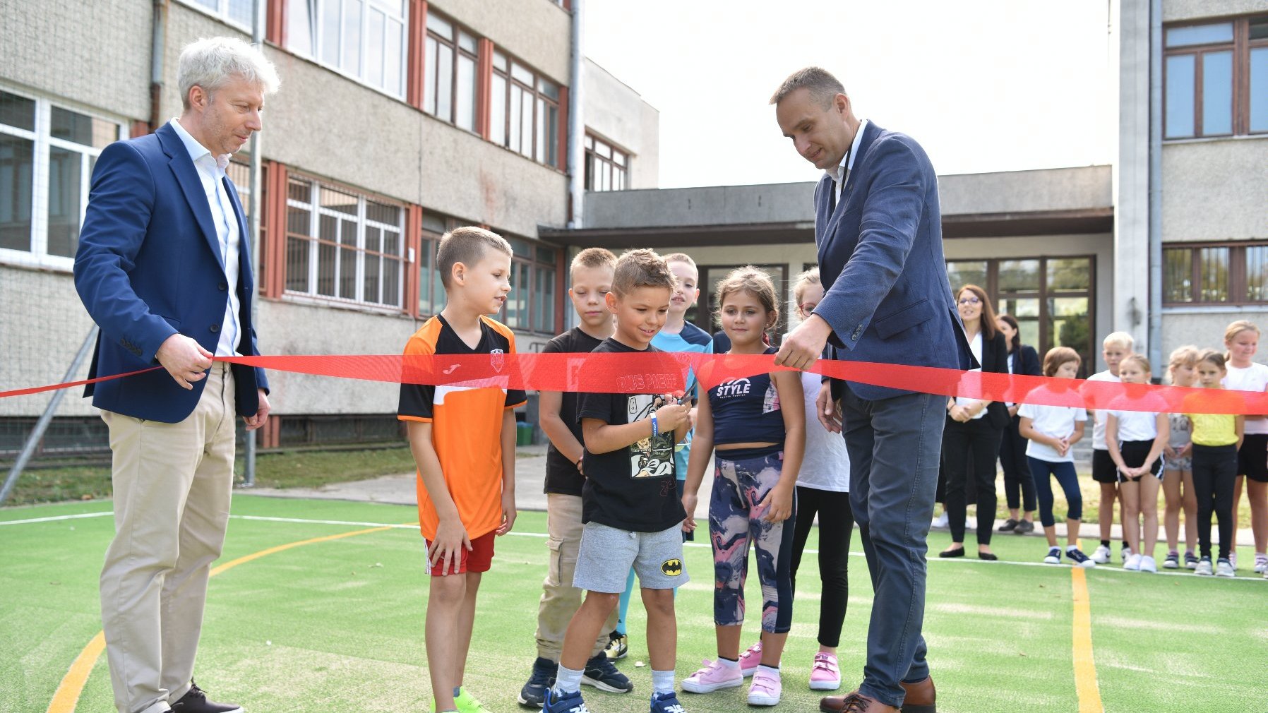 Zdjęcie przedstawia przecinanie wstęgi na nowym boisku. Są na nim dzieci i dorośli.