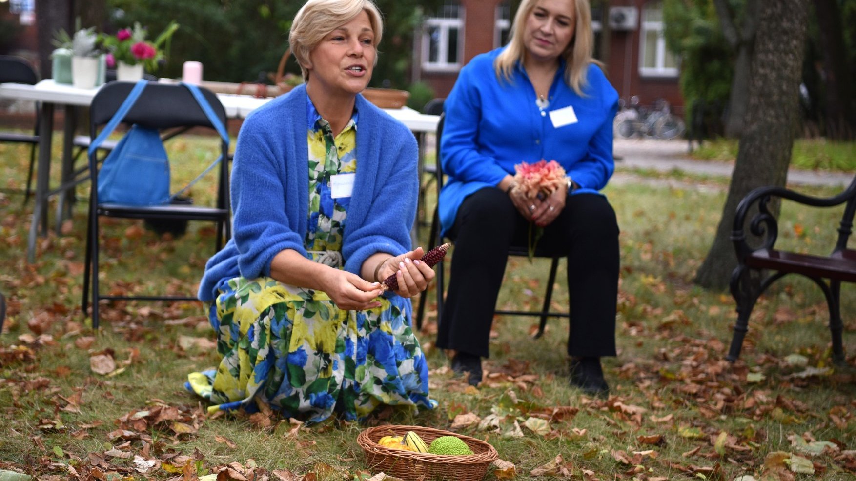 Zdjęcie przedstawia dwie kobiety, jedna kuca, druga siedzi na krześle. Na ziemi leży koszyk z owocami.