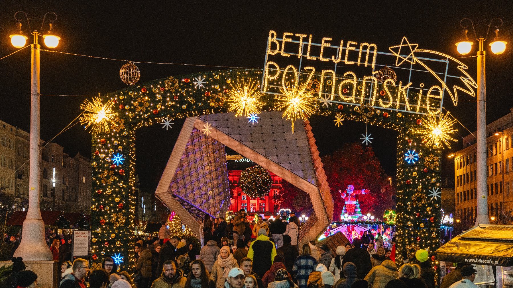 Zdjęcie przedstawia tłum ludzi na placu Wolności. Widać na nim m.in. fontannę i świecący neon z napisem Betlejem Poznańskie.