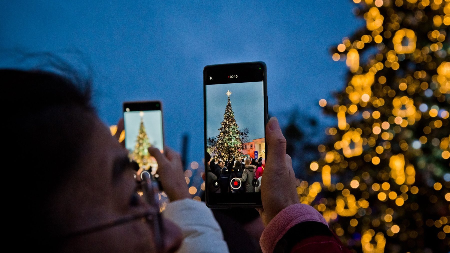 Zdjęcie przedstawia ludzi robiących zdjęcia choince. Rozświetlone drzewko widać na ekranach telefonów.