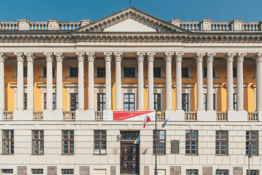 "Budynek w stylu klasycznym z symetryczną fasadą. Na fasadzie widoczne są wysokie, ozdobne kolumny podtrzymujące fronton z bogatymi detalami. Budynek jest pomalowany na żółto i biało, z dużymi oknami i dekoracyjnymi elementami. Na przodzie budynku wisi czerwono-biały baner, a przy wejściu widoczne są dwie flagi. Budynek wydaje się być ważnym lub historycznym obiektem, prawdopodobnie instytucją rządową lub kulturalną. - grafika artykułu