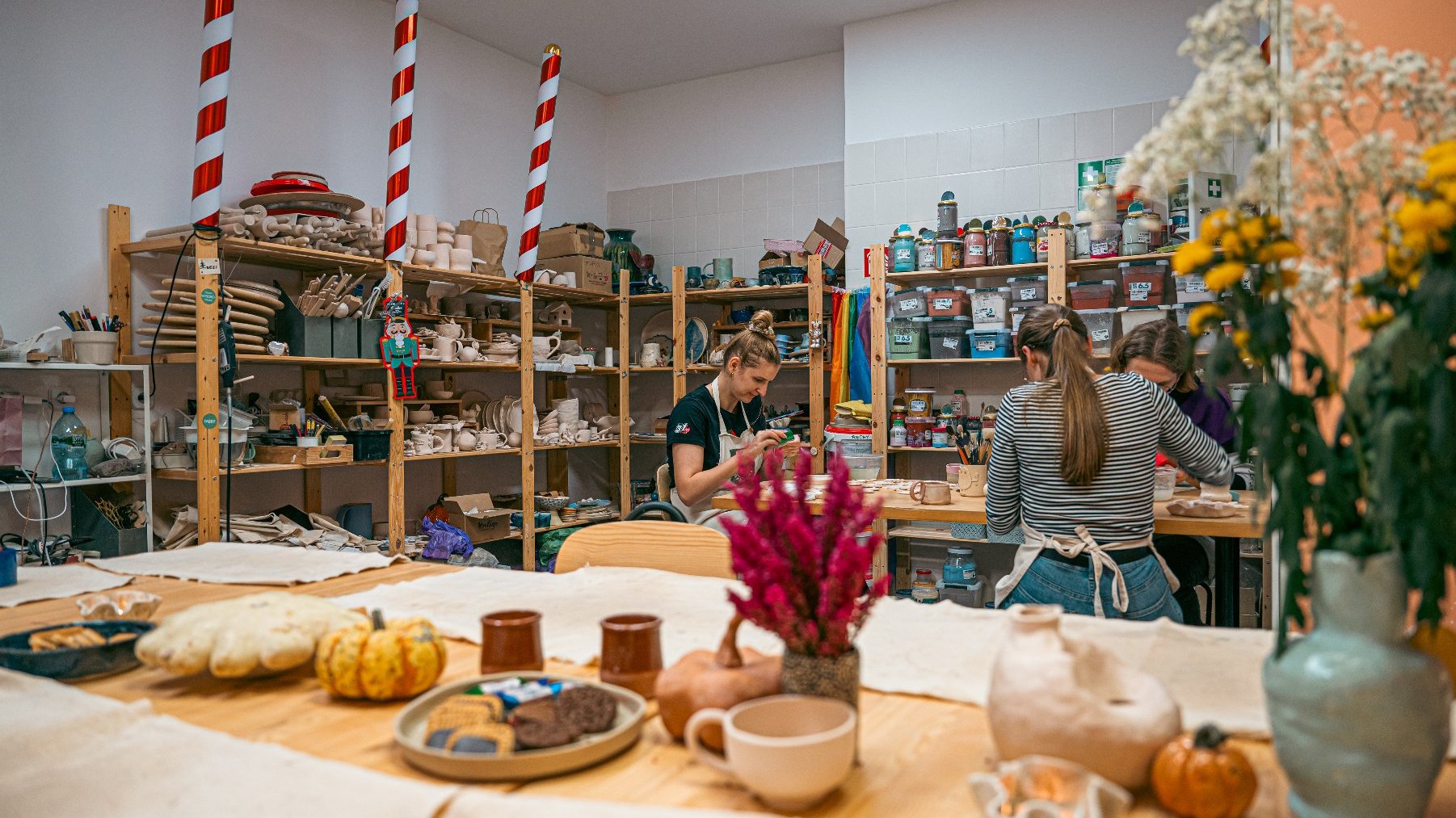 Na zdjęciu widać warsztaty ceramiczne. Uczestnicy skupiają się na swoich zadaniach. Półki są zaopatrzone w różnorodne wyroby ceramiczne i narzędzia. Widoczne są małe dynie oraz wazon z kwiatami.