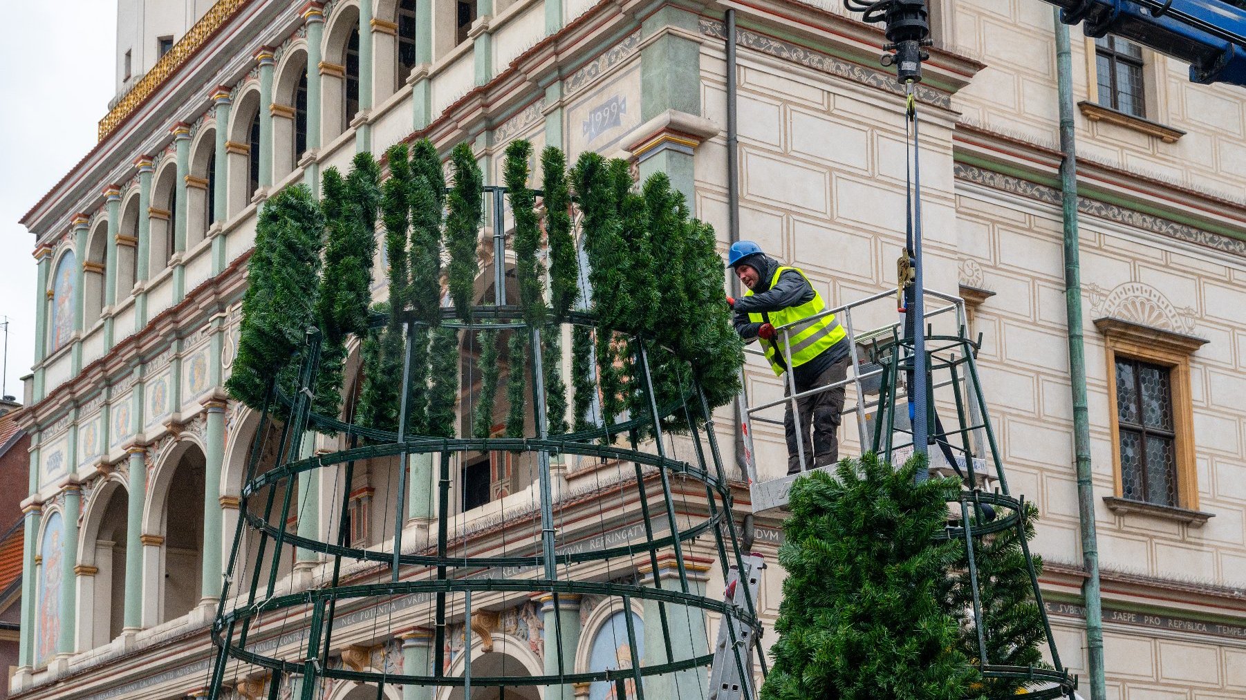 Osoba pracująca przy składaniu dużej sztucznej choinki. Stoi na platformie, przymocowując zielone gałązki do metalowej ramy w kształcie choinki. W tle widać zabytkowy budynek z ozdobnymi detalami architektonicznymi, takimi jak łuki i elementy dekoracyjne. Scena wygląda na część przygotowań do świątecznego pokazu publicznego. Użycie dźwigu i sprzętu ochronnego wskazuje na profesjonalną instalację.