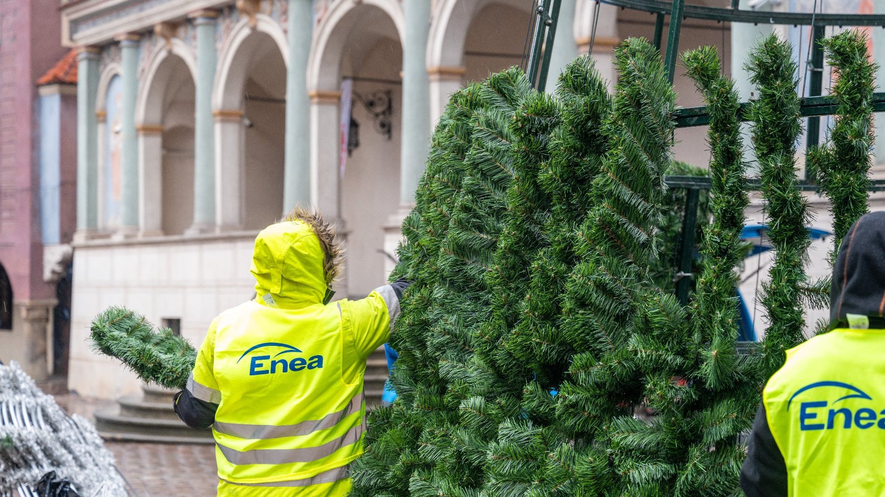 Na zdjęciu znajdują się dwie osoby ubrane w jaskrawe, żółte kurtki z logo 'Enea' na plecach. Wydają się montować lub dekorować dużą sztuczną choinkę. Choinka jest zbudowana z zielonych sztucznych gałęzi przymocowanych do metalowej ramy. W tle widoczny jest budynek z łukowymi kolumnami i ozdobnymi detalami architektonicznymi, co sugeruje, że znajduje się on w publicznej lub historycznej okolicy.