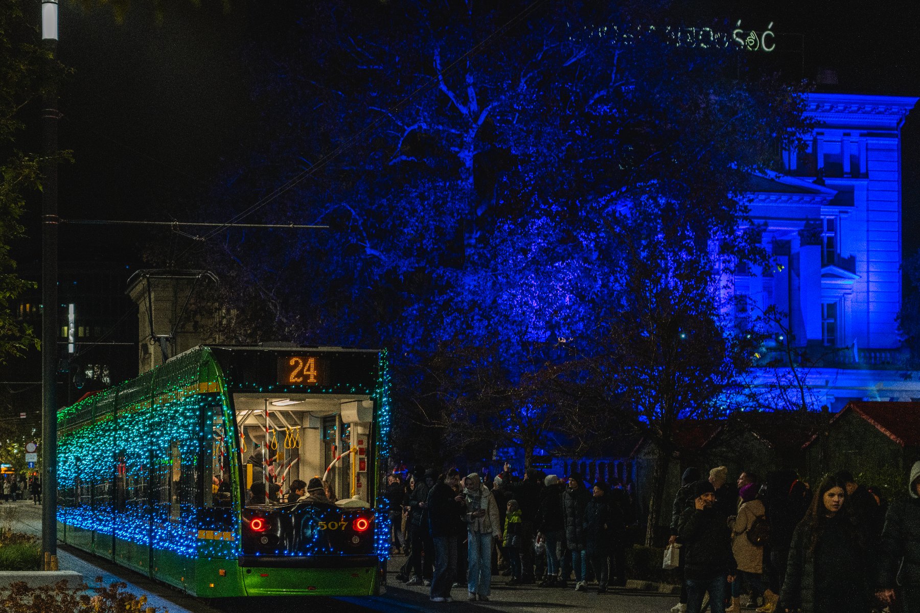 Tramwaj z numerem 24, ozdobiony niebieskimi i zielonymi światłami, co nadaje mu świąteczny wygląd. Wewnątrz tramwaju znajdują się ludzie, a na zewnątrz zgromadził się tłum, prawdopodobnie czekający na wejście lub obserwujący tramwaj. W tle widać budynek oświetlony niebieskimi światłami oraz drzewa. - grafika artykułu