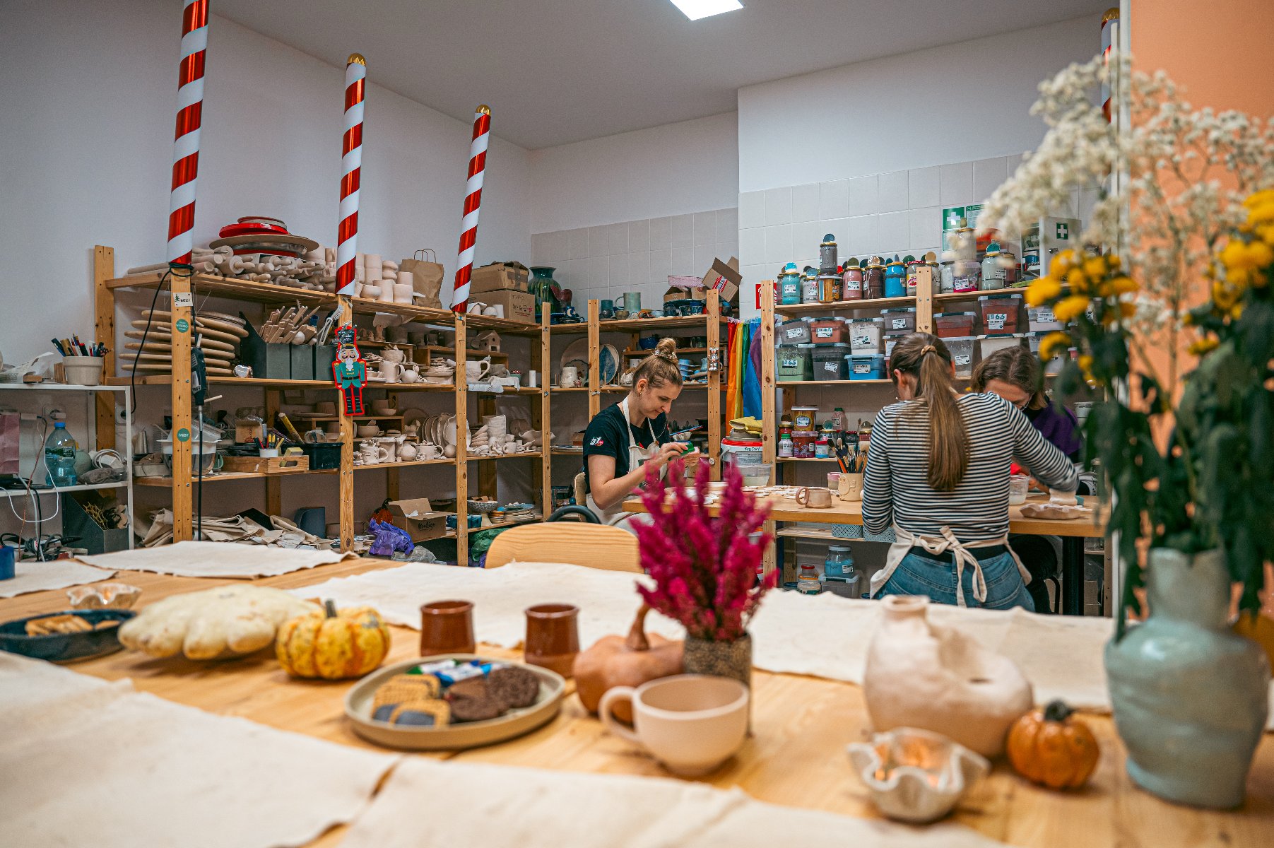 Na zdjęciu widać warsztaty ceramiczne. Uczestnicy skupiają się na swoich zadaniach. Półki są zaopatrzone w różnorodne wyroby ceramiczne i narzędzia. Widoczne są małe dynie oraz wazon z kwiatami. - grafika artykułu