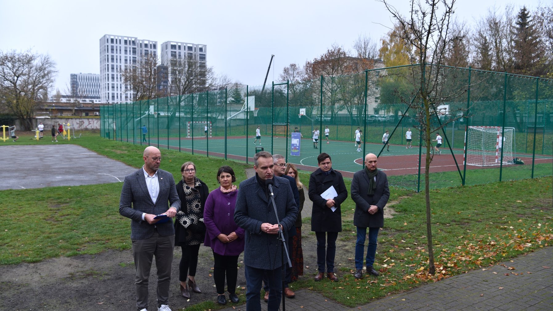 Zdjęcie przedstawia uczestników konferencji prasowej. Do mikrofonu mówi mężczyzna. Za ich plecami znajduje się boisko.