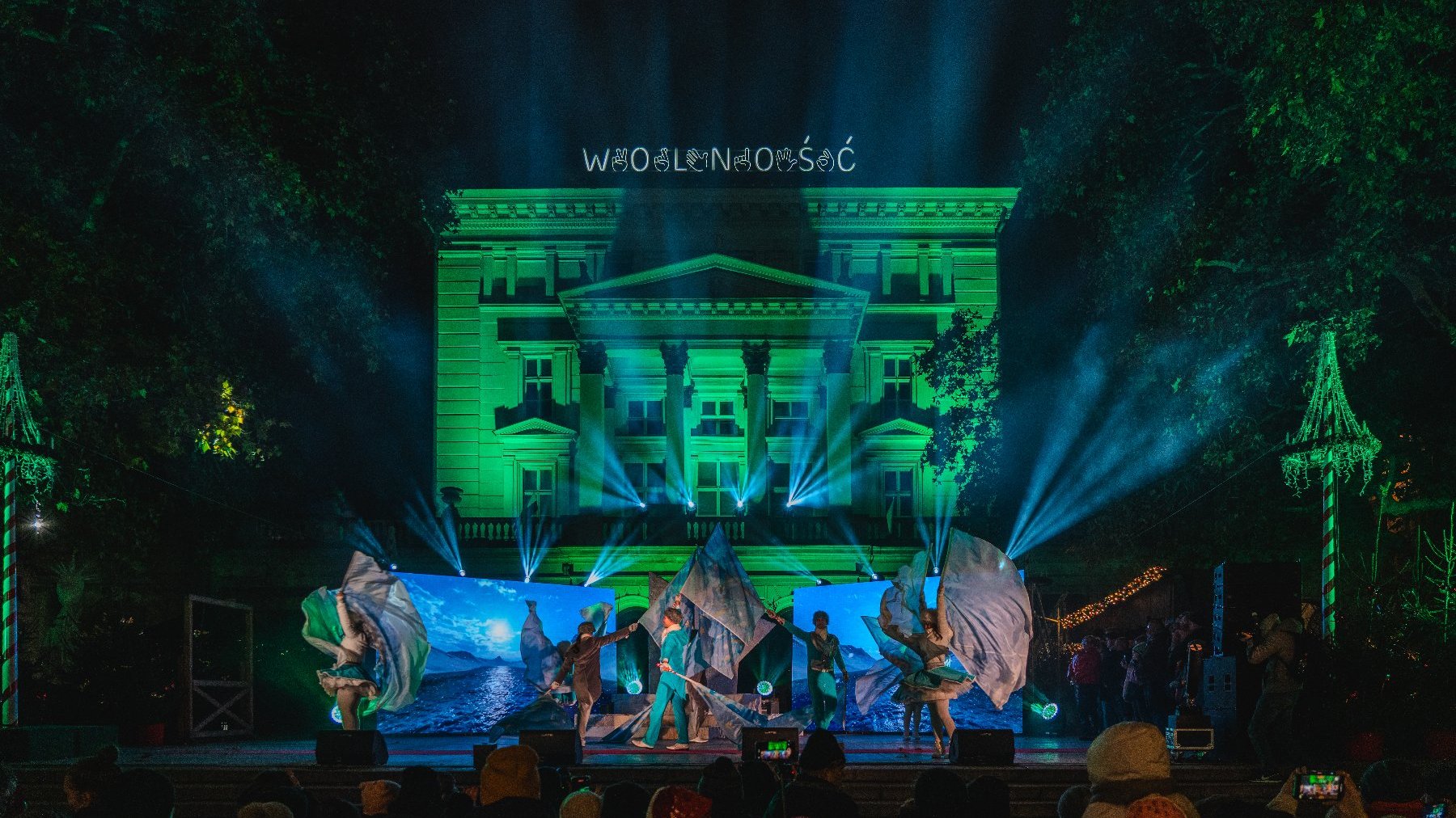 Zdjęcie przedstawia ludzi tańczących na scenie i podświetlony budynek.