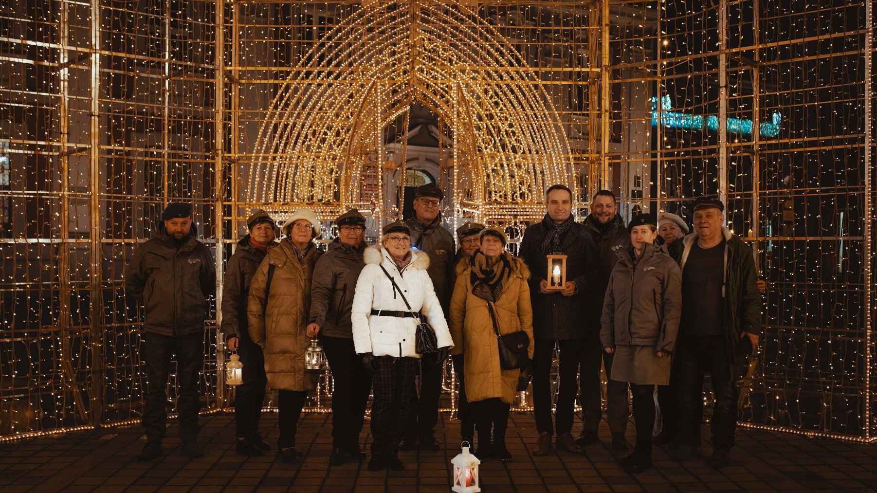 Grupa ludzi stoi razem przed dużą, oświetloną strukturą. Struktura jest udekorowana licznymi małymi światełkami, tworzącymi łuk i kratowy wzór. Niektóre osoby w grupie trzymają latarnie, a jedna latarnia jest postawiona na ziemi przed nimi. Scena wygląda na świąteczną, prawdopodobnie związaną ze świętem lub celebracją, biorąc pod uwagę dekoracyjne światła i latarnie.