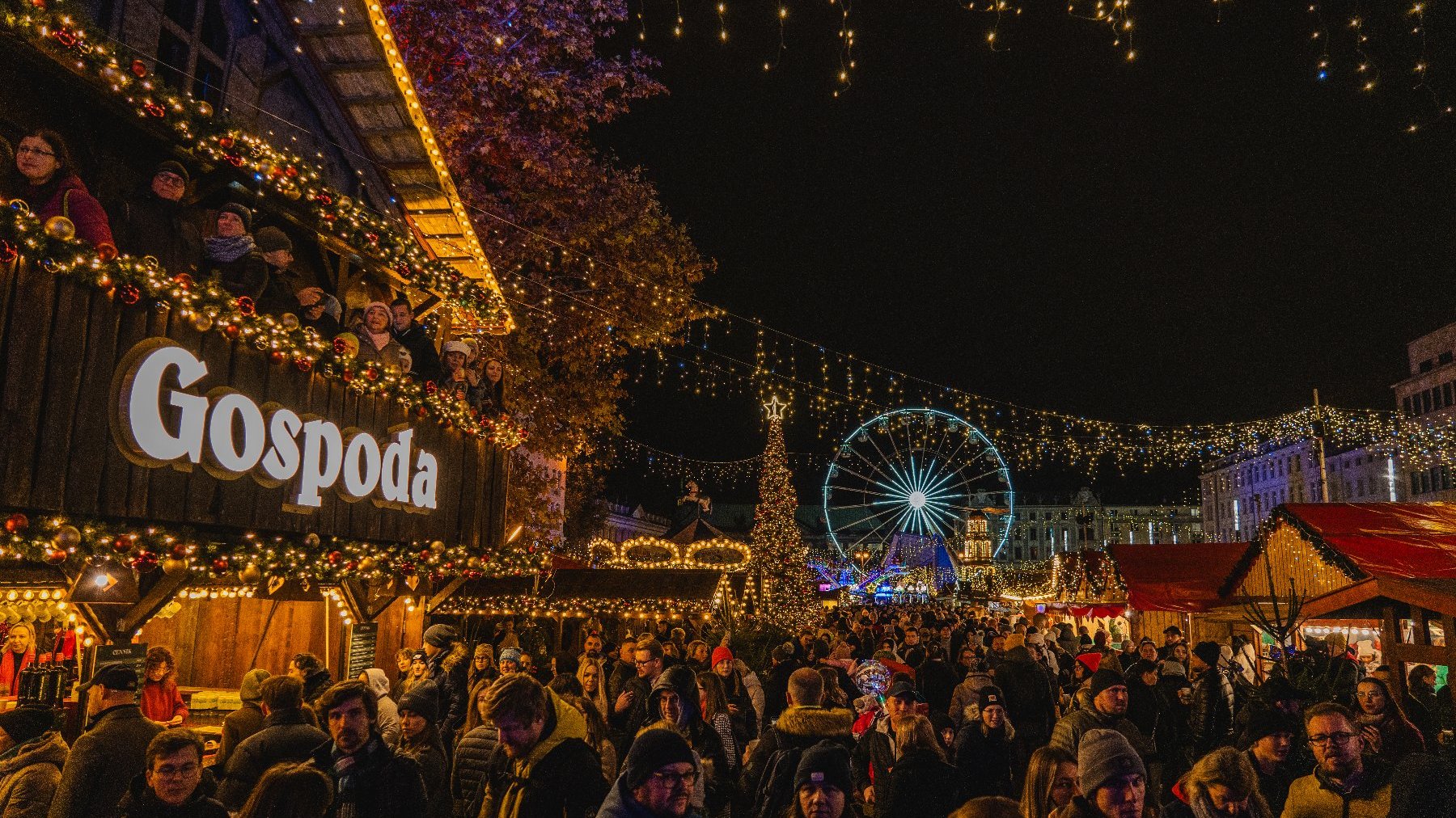 Zdjęcie przedstawia tłum ludzi, drewniany domek z napisem Gospoda i inne domki handlowe, młyńkie koło, iluminacje świąteczne i choinkę.