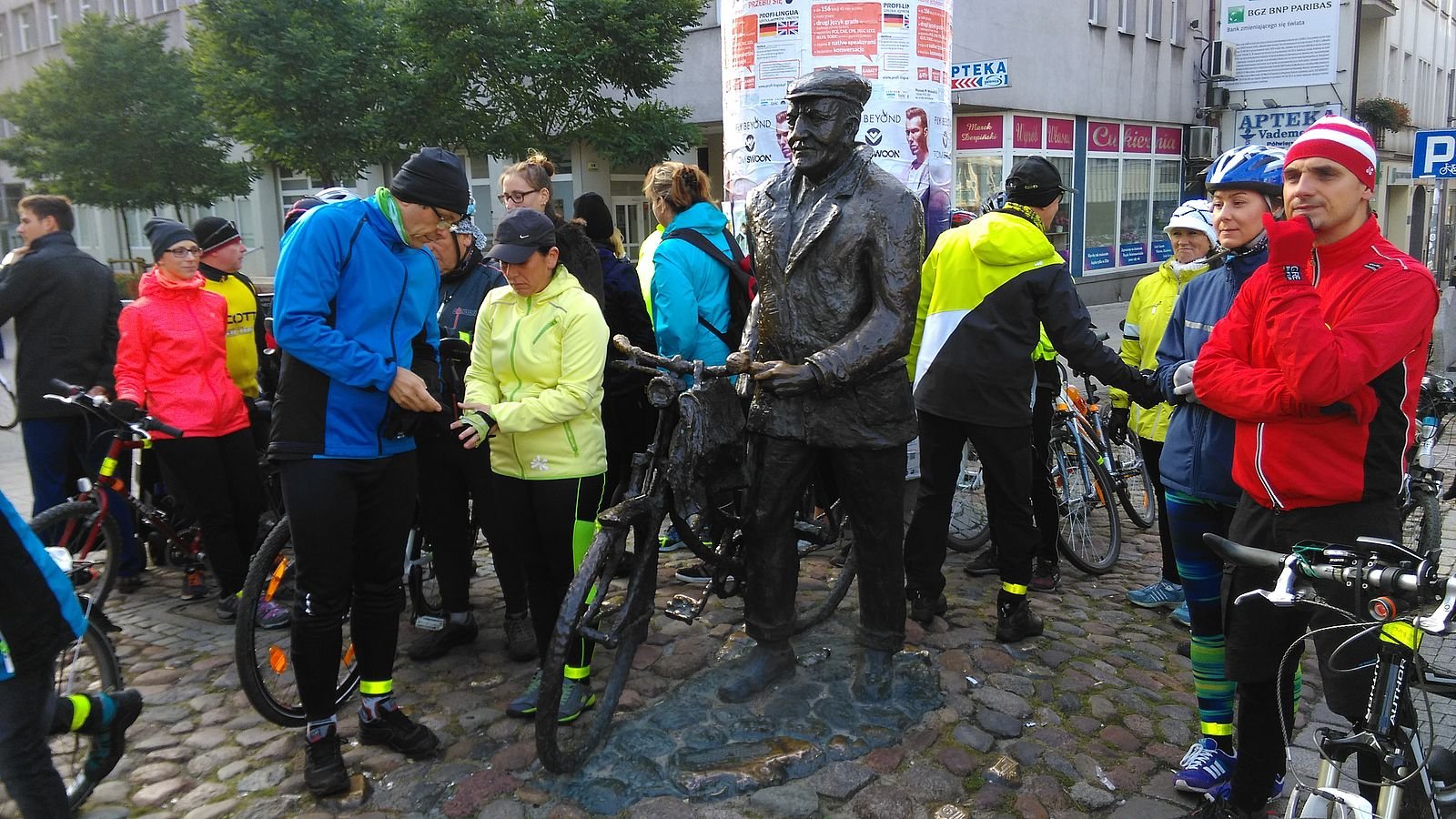 Zdjęcie, grupa rowerzystów przygotowuje się do startu w rajdzie. Są kolorowo ubrani, na głowach mają czapki i kaski rowerowe.