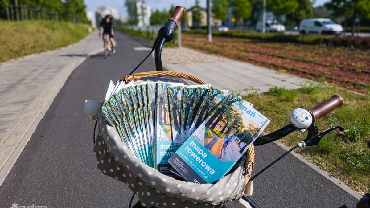 Zdjęcie, rower stoi na ścieżce rowerowej. W koszyku zawieszonym na kierownicy znajduje się plik map rowerowych Poznania.