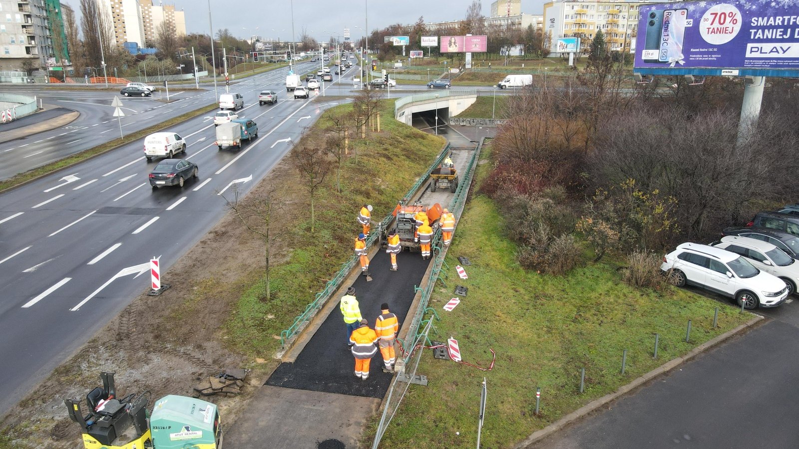 Roboty drogowe zwiazane z trasą rowerową przy przystanku PST Słowniańska. Pracownicy drogowi ubrani w stroje odblaskowe, maszyna do układania nawierzchni, obok skrzyżowanie drogowe.