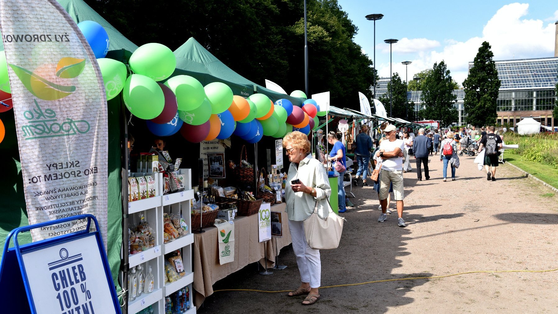 Zdjęcie przedstawia stoiska z produktami w parku oraz spacerujących alejkami ludzi.