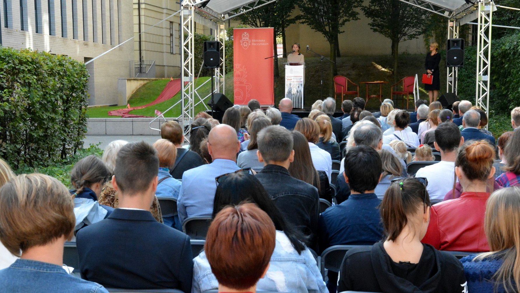 Zdjęcie przedstawia scenę, na której kobieta przemawia do mikrofonu oraz słuchających ją ludzi, siedzących na krzesłach.