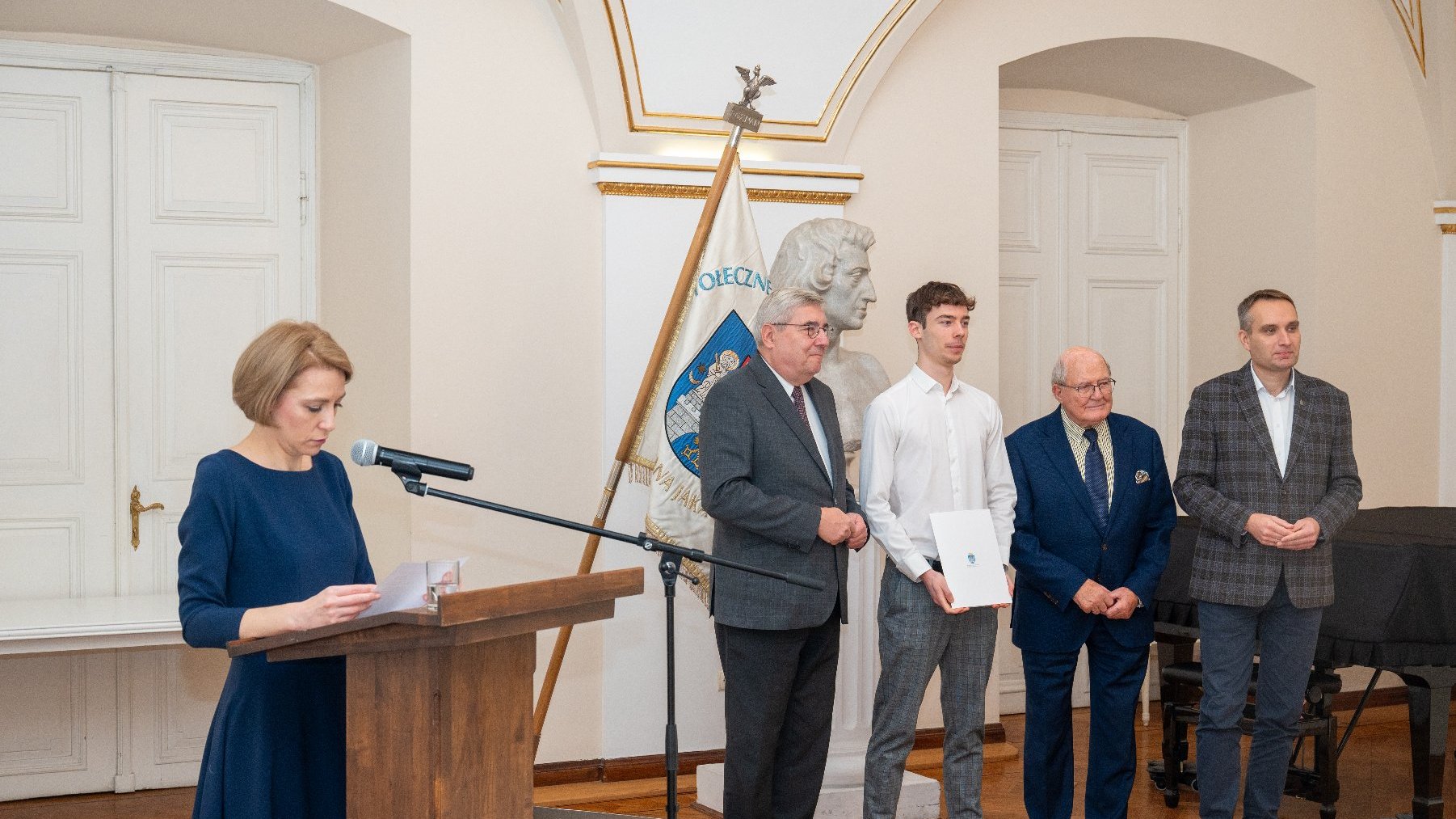 Zdjęcie przedstawia trójkę mężczyzn i młodego chłopaka pozujących wspólnie do zdjęcia oraz kobietę przy mównicy.