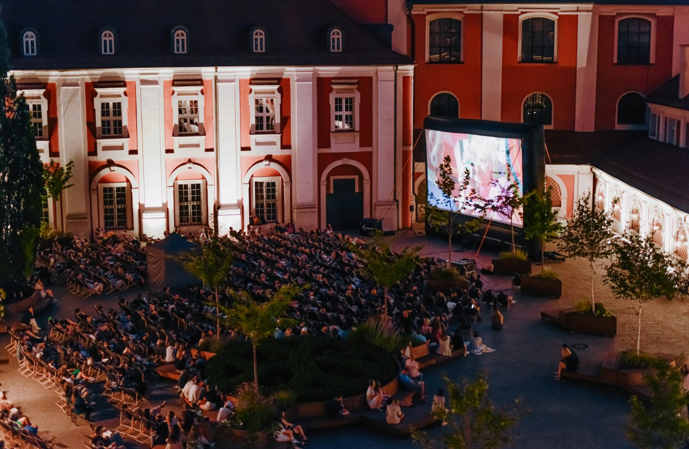 Zdjęcie przedstawia ludzi na leżakach oglądających film na telebimie na dziedzińcu urzędu miasta. - grafika artykułu