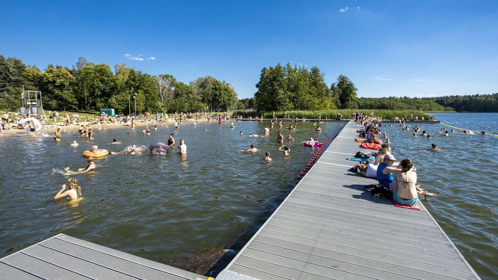 Zdjęcie przedstawia ludzi w wodzie, na pomoście i na plaży.