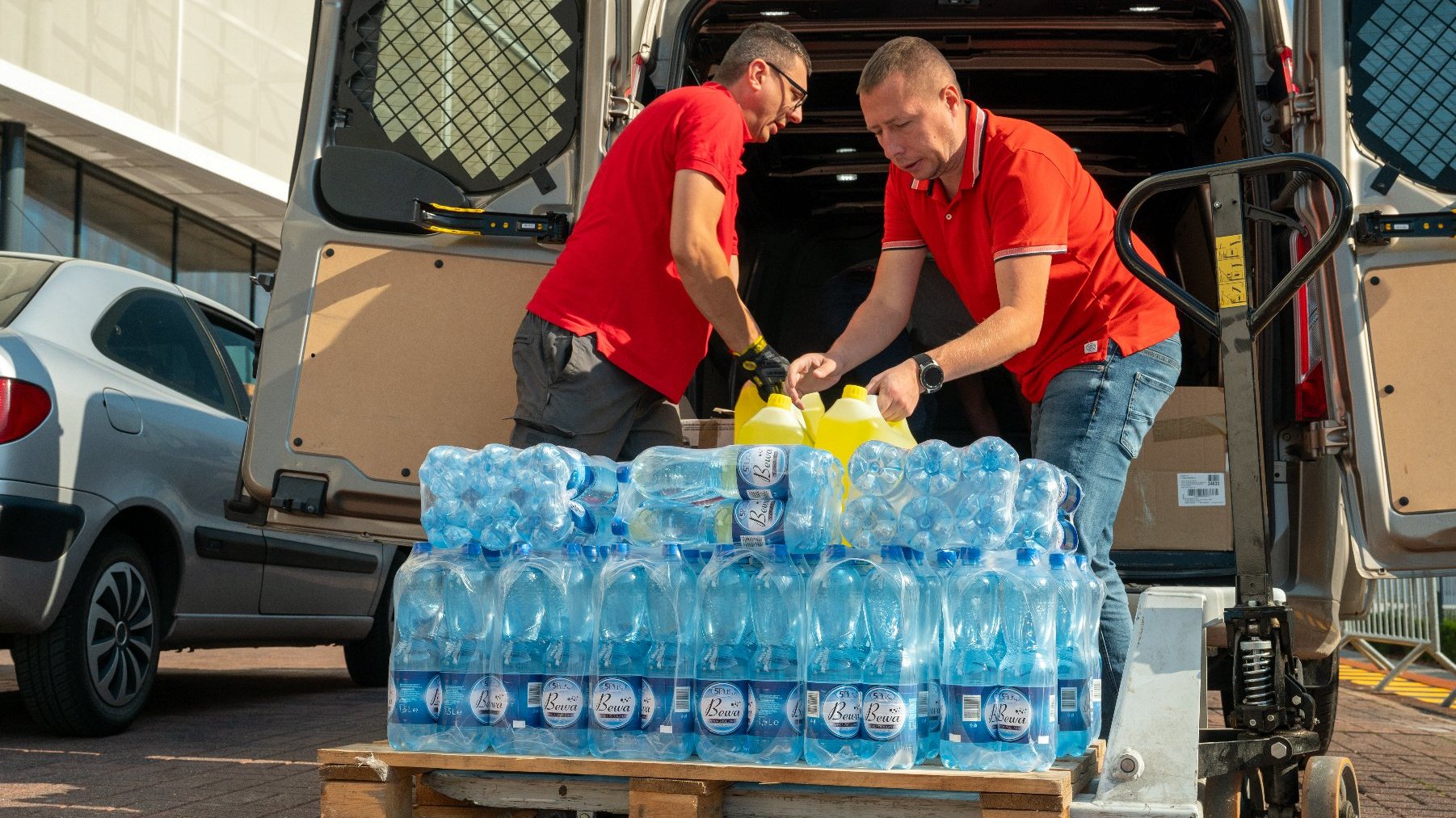 Na zdjęciu dwóch mężczyzn wypakowujących zgrzewki butelkowanej wody