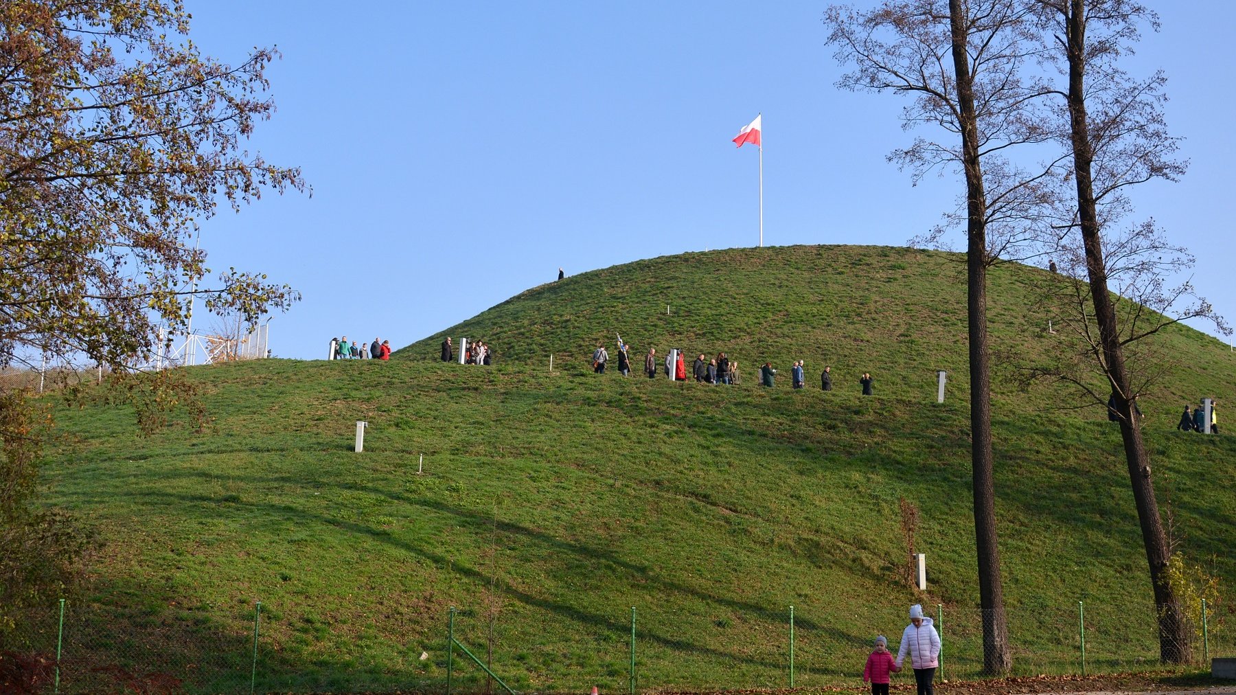 Zdjęcie przedstawia Kopiec Wolności i zwiedzajacych go ludzi. - grafika artykułu