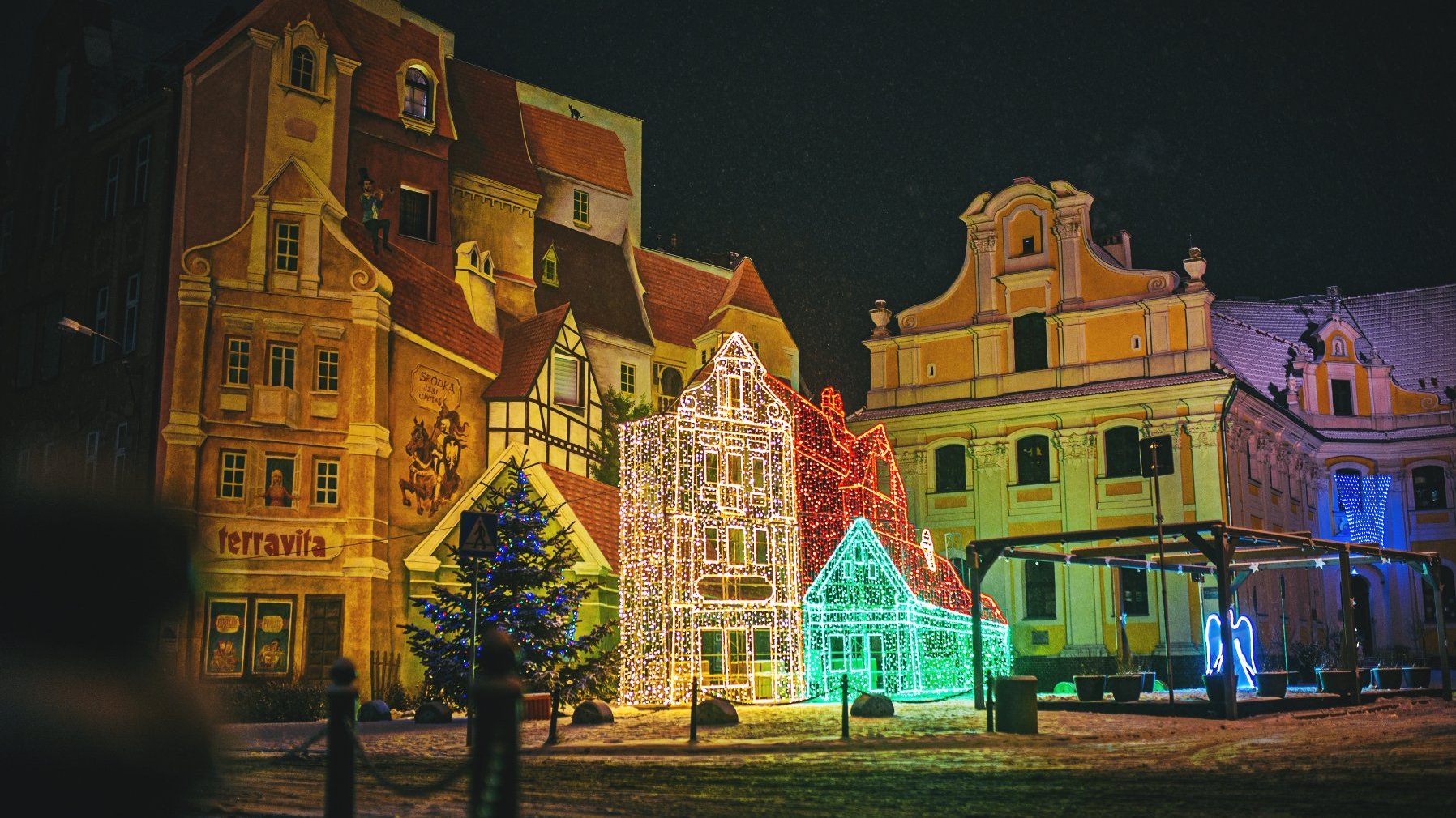 Zdjęcie przedstawia budynki. Na jednym z nich znajduje się mural. Widać też iluminację w kształcie budynków.