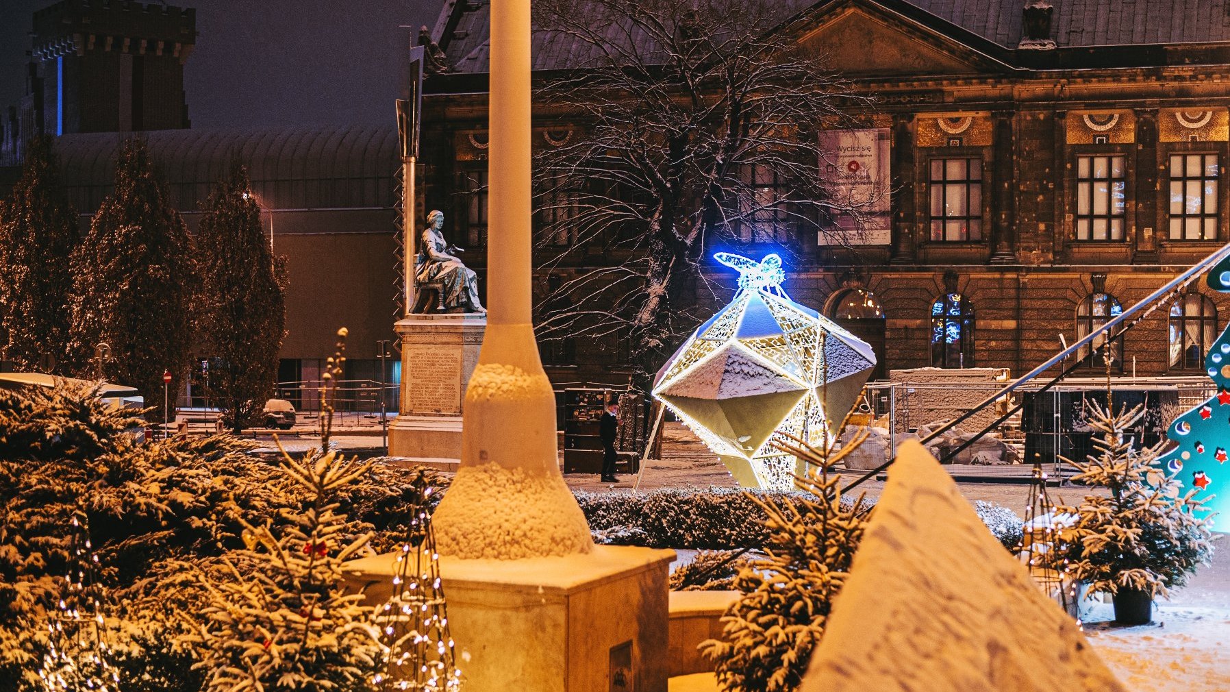 Zdjęcie przedstawia iluminację w kształcie diamentu, na którym siedzi mucha, a także budynki, pokryte śniegiem krzewy i pomnik.