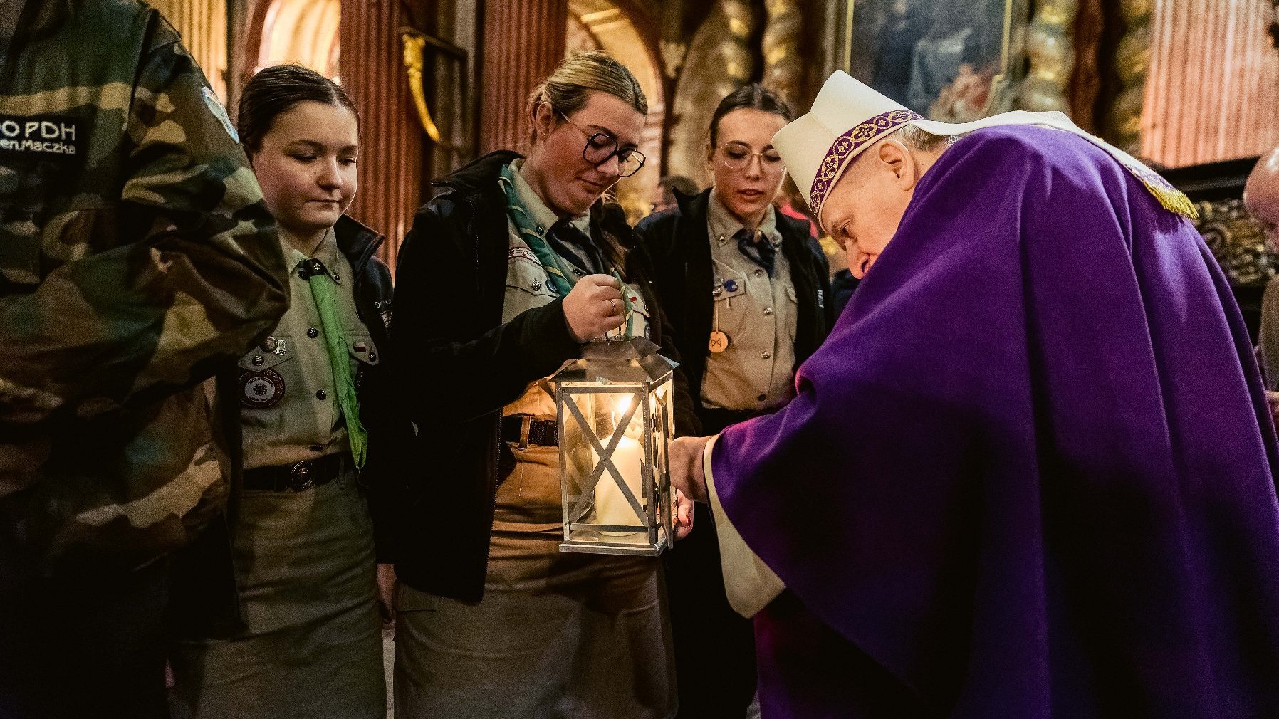 Grupa ludzi, niektórzy ubrani w uniformy przypominające mundury harcerskie, oraz jedna osoba ubrana w fioletową szatę typową dla duchownych. Osoba w fioletowej szacie wydaje się zapalać lub błogosławić latarnię trzymaną przez jedną z uniformowanych osób. Scena ma miejsce wewnątrz kościoła lub podobnego budynku sakralnego, co wskazują ozdobne kolumny i religijne dzieła sztuki w tle.